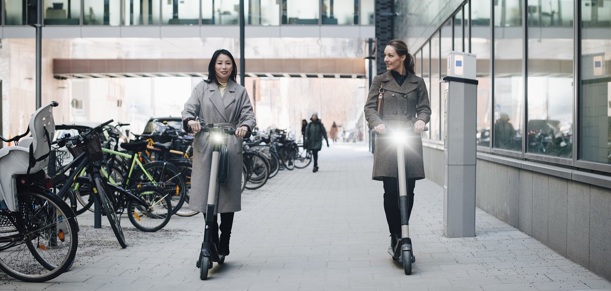 Två kvinnor åker på varsin elsparkcykel i stadsmiljö