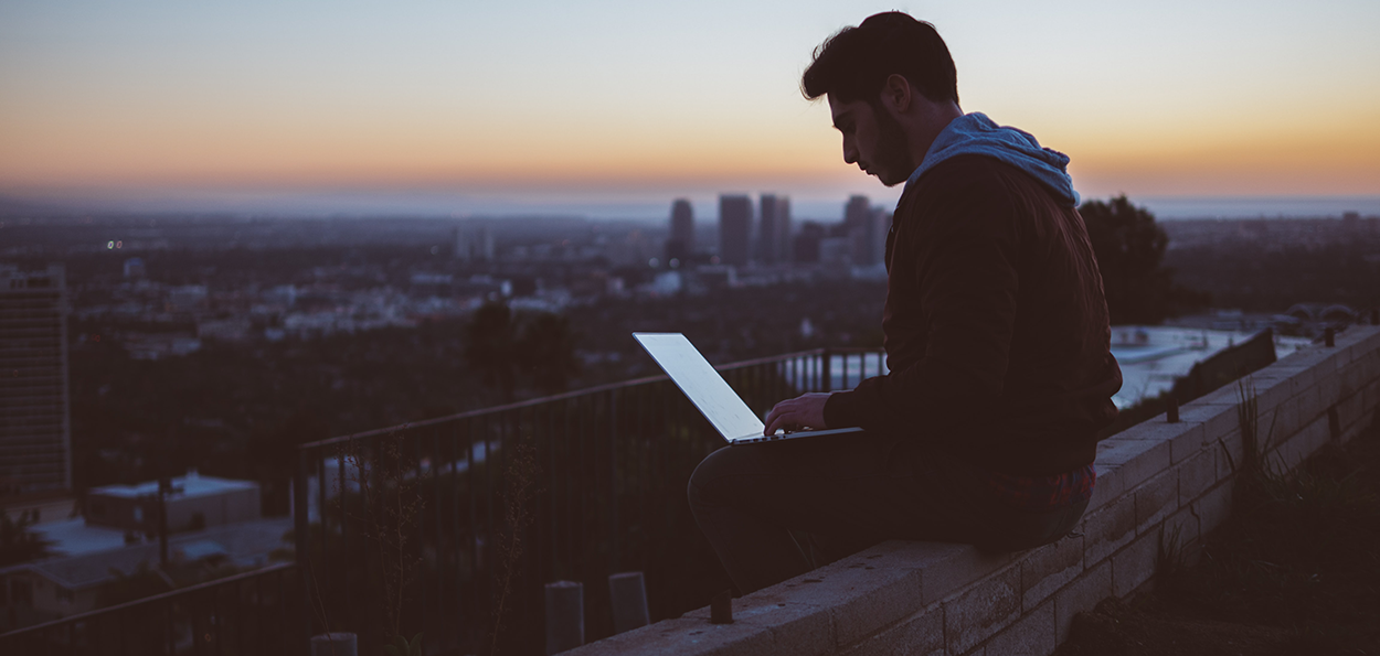 Ung man arbetar vid laptop utomhus i skymning med utsikt över stadsmiljö